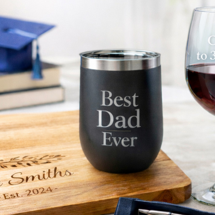 Personalized drinkware and cutting board displayed on a light marble surface, featuring a tumbler, wine glass with frosted engraving, and engraved bamboo cutting board