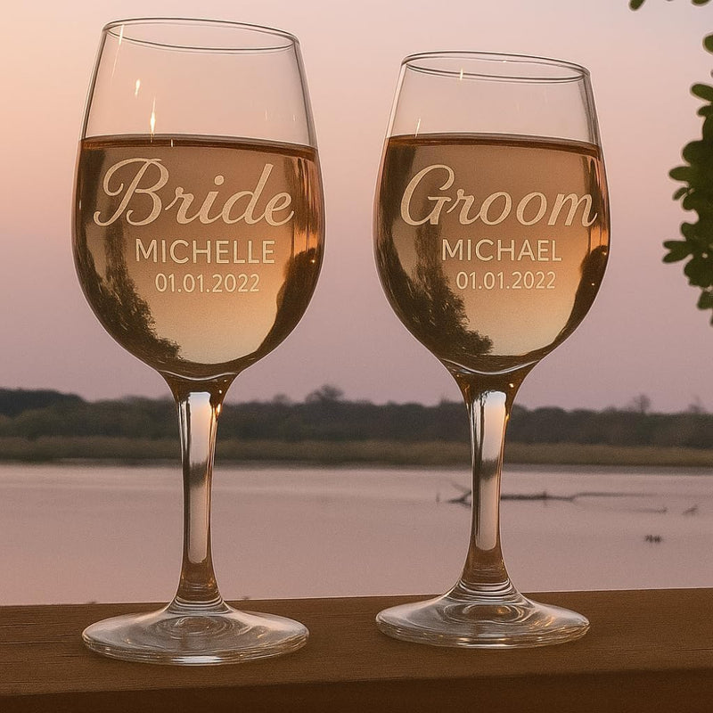 Two wine glasses with 'Bride' and 'Groom' engraved names and dates, filled with a golden liquid, against a sunset or sunrise backdrop.
