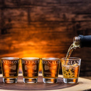 Four shot glasses with engraved names being filled with a liquid on a wooden surface.