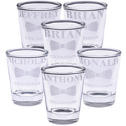 Personalized groomsmen shot glasses with engraved names, bow tie design, wedding role, and date arranged in a group on a white background