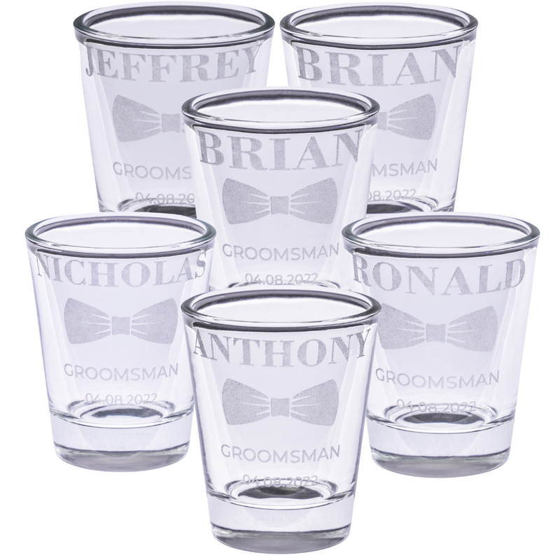 Personalized groomsmen shot glasses with engraved names, bow tie design, wedding role, and date arranged in a group on a white background