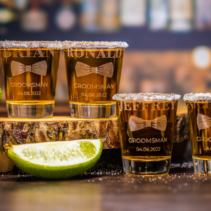 Personalized groomsman shot glasses engraved with names, bow tie design, and wedding date, filled with whiskey and salt rim